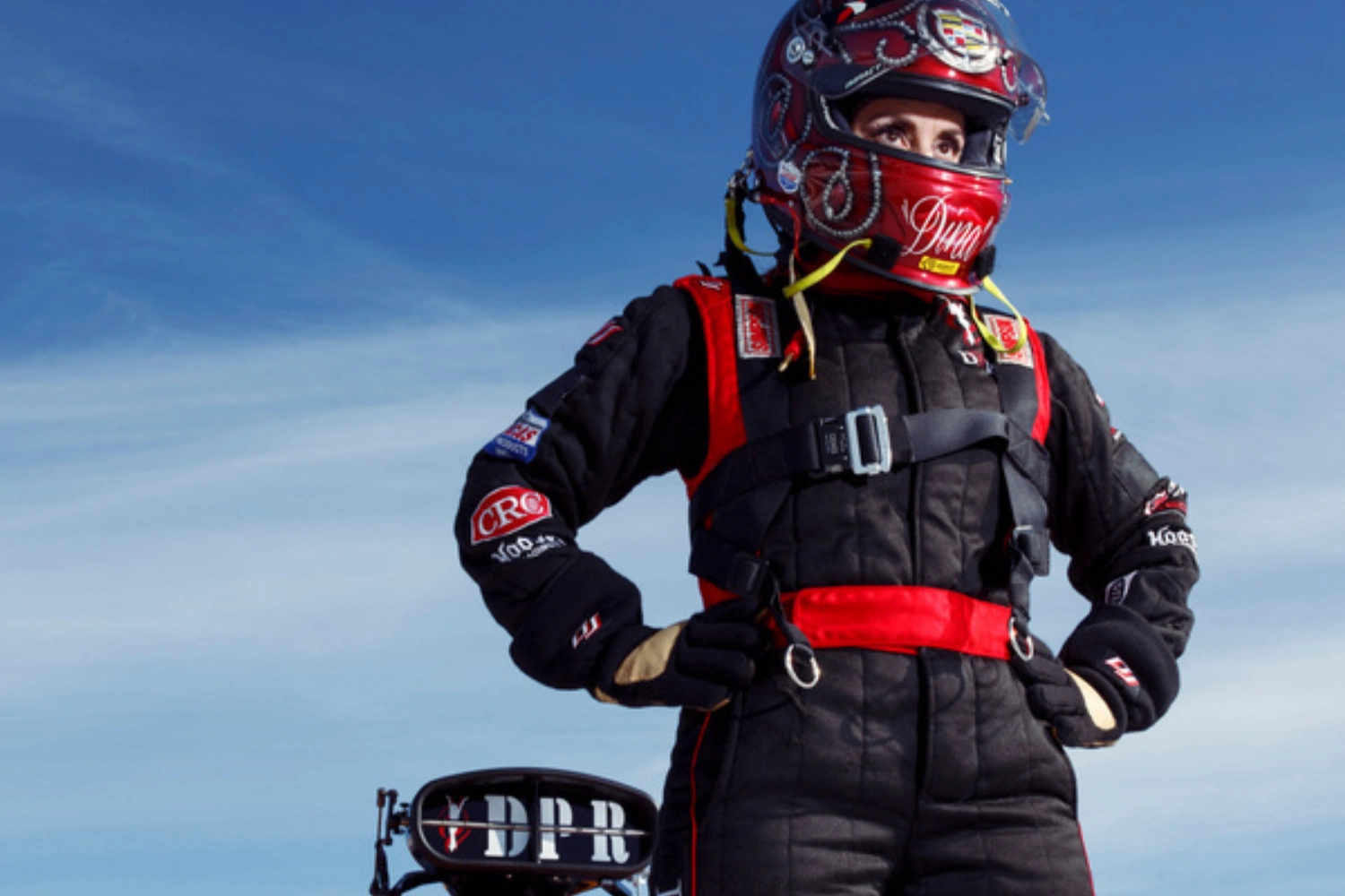 Dina Parise in her red and black fire suit and helmet, hands on hips, against a blue sky